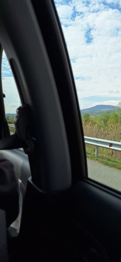 The image shows a view from inside a car, possibly a sunroof perspective. Outside the car, there is a roadside with a guardrail, vegetation, and a distant mountain. The sky is visible with clouds. There is no indication of being in the city of Valkovo, Bulgaria. There are no visible people.