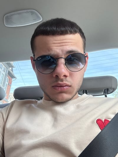 A medium shot shows a man inside a vehicle. He is wearing round, dark-tinted sunglasses with the Ray-Ban logo visible on the left lens. He has dark hair, neatly trimmed, and a light beard. He is wearing a light-colored sweatshirt with a red clover or flower design on the upper right. A dark gray seatbelt is diagonally across his chest. Behind him, the vehicle's interior is visible, including a light gray headrest, a ceiling light, and the car window showing a view of a building with red roof tiles. The building's architecture suggests an urban environment. However, it's impossible to verify this is Cochem, Germany. The perspective is angled, as if the photo was taken as a selfie with the camera rotated.