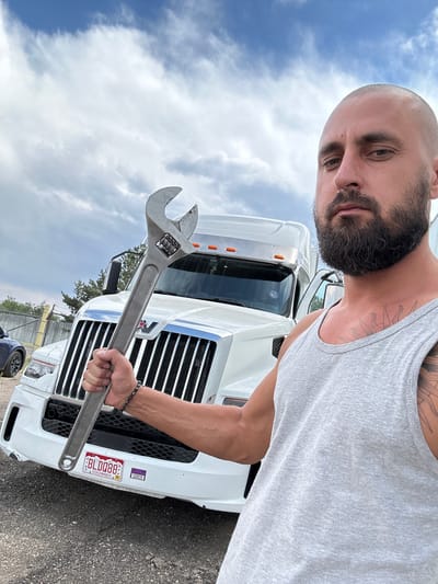 A bald, bearded Caucasian man in a grey tank top holds a large adjustable wrench in his right hand, raised in front of a white semi-truck. He is looking at the camera with a serious expression. The truck's grille and headlights are visible, as is a Colorado license plate that reads "BLDQ88." The man has tattoos on his chest and arm. The scene appears to be outdoors, with a cloudy sky overhead and gravel on the ground. A chain-link fence and trees are visible in the background. This image was taken in Aurora, Colorado.
