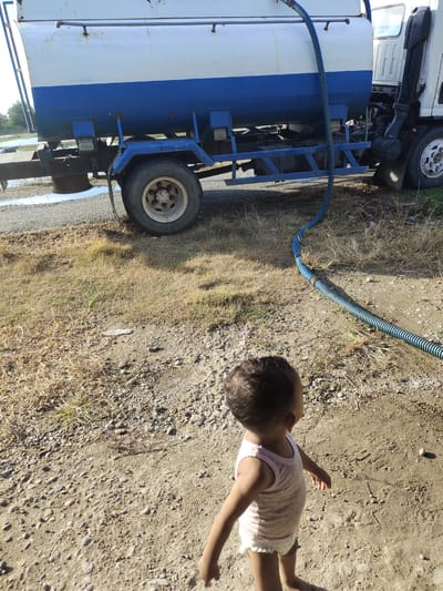In Teupinkuyun, Indonesia, a child stands on a sandy and grassy ground near a water truck. The truck has a white and blue tank and a blue hose connected to it, running across the ground. The child is wearing a light pink tank top and a diaper, and is facing away from the camera, looking towards the truck. The environment appears to be outdoors, possibly a rural or undeveloped area, with dry grass and scattered stones on the ground.