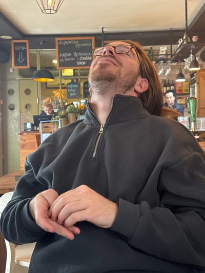 The image depicts an indoor scene within a casual dining or cafe establishment located in Raska Municipality, Serbia. In the foreground, a light-skinned male individual with a beard and long brown hair is seated at a wooden table. He wears eyeglasses with brown frames and a dark grey fleece pullover with a silver zipper partially open at the collar. His head is tilted sharply backward, with his chin elevated, and his eyes appear closed or directed toward the ceiling. His hands are clasped or interlocked in front of him on the table.

The background shows the interior of the establishment. To the left, a black chalkboard sign displays "KASA" (Cash Register) and a downward arrow in white text. Adjacent to it, another chalkboard lists menu items in Serbian, including "PUNJENA PLJESKAVICA SA ŠUNKOM I CHEDDAR SIROM" (Stuffed Pljeskavica with Ham and Cheddar Cheese). Behind a wooden counter on the left, a female individual with reddish-blonde hair in a bun is observed looking down, presumably interacting with a point-of-sale system. Further to the right and deeper in the scene, a male individual with dark hair is partially visible behind a bar area, looking right. The environment includes various lighting fixtures, such as a decorative ceiling light and a black dome-shaped lamp with a gold interior, along with a green beer tap, bottles, glasses, and potted plants on counters. Metallic circular wall elements and a snowflake-shaped decoration are also present.