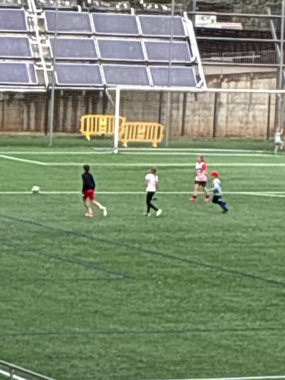 The image shows a soccer field with four people playing soccer. The field is green with white lines. A soccer ball is on the ground near one of the players. To the left of the field is a building with barred windows. The photo was taken in Esplugues de Llobregat, Spain.