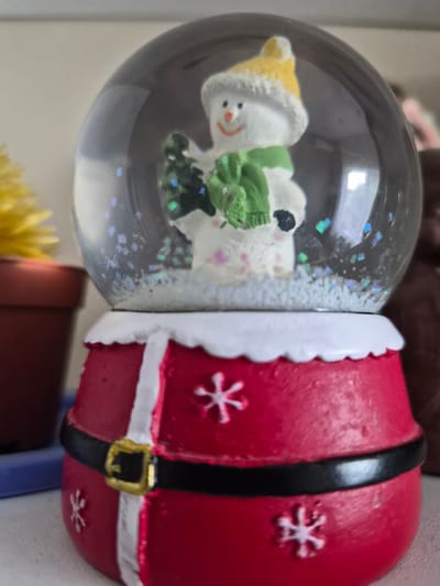 This image shows a close-up of a Christmas-themed snow globe. The snow globe is partially submerged in water and contains a small snowman wearing a yellow hat and a green scarf. The snowman has a painted smile and stick-figure eyes and nose. The snow globe is filled with glitter that appears to be shimmering. The base of the snow globe is red and designed to look like the suit of Santa Claus, complete with a black belt and a golden buckle, and white trim. The snow globe is resting on a surface, possibly a shelf or table, and a blurred yellow object, possibly a flower, is visible in the lower part of the image. The lighting suggests it is indoors, and there are no obvious cues to indicate the time of day or weather. The context provided is "Lenina, Russia," which suggests a possible location but offers no specific environmental details visible in the image itself. No text is visible in the image.