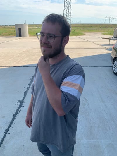 A man with light skin, brown hair, and a beard stands on a paved surface under a cloudy sky. He is wearing glasses, a gray t-shirt with horizontal peach and white stripes on the sleeves, and blue jeans. He has his right hand on his chin and is looking to his left. In the background, there are industrial structures, including a tall metal tower and power lines, as well as a small concrete building. The scene appears to be an open, somewhat industrial area, possibly on the outskirts of the city of Pivdennoukrainsk, Ukraine, given the presence of large power infrastructure. A portion of a dark-colored car is visible on the right side of the frame.