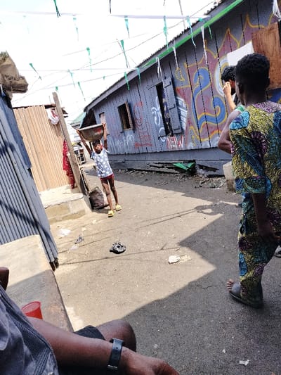 The scene is set on a narrow, unpaved pathway within Oworonshoki, Nigeria, under bright daylight. Overhead, multiple strings with green and white triangular pennants are strung across the street.
To the right, a single-story structure with grey wooden plank walls displays extensive multi-colored graffiti, including blue, yellow, and red abstract designs, along with white text fragments such as "NEW" and "CREW." A shuttered window is visible on this building. To the left, another structure made of vertical wooden slats or corrugated metal is partially visible. The pathway itself is a mix of dirt and asphalt, with scattered debris including plastic bags and other refuse.
Three individuals are present in the midground. In the center-left, a child, approximately 7-10 years old, with light skin, wears a white t-shirt with dark star patterns and red shorts. This child is actively balancing a grey metallic basin on their head, held with both raised hands, and wears yellow open-toed sandals. To the right, an adult or older youth, with dark skin, stands with their back largely to the viewer, dressed in a vibrant, multi-colored patterned shirt and matching pants featuring yellow, blue, green, and black designs. They wear grey open-toed sandals. Partially obscured behind this individual is another person with dark, curly hair, facing towards the graffiti-covered building.
In the extreme lower-left foreground, a portion of an arm wearing a black wristwatch and a dark grey shirt is visible, alongside a red plastic cup. Shadows are cast across the pathway.