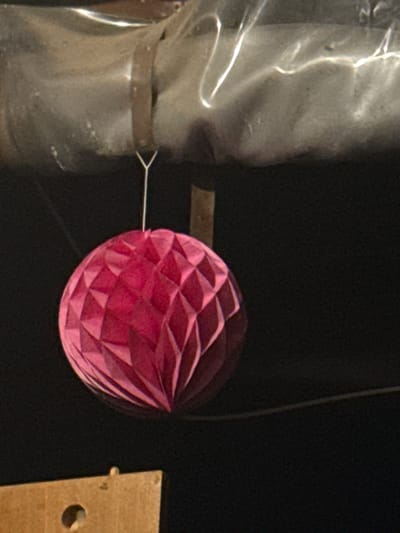 The image shows a close-up of a pink, honeycomb-shaped paper lantern. The lantern is suspended by a thin white string, which is attached to a metallic band around a cylindrical object on the left. This object appears to be made of a translucent, crinkled material, possibly plastic wrap or a similar flexible substance, and has a somewhat metallic sheen. A dark, coarse-textured band encircles this object. To the right of the lantern, a portion of a light brown, rectangular object with a circular hole is visible. This object is made of a coarse material, possibly wood or thick cardboard.

The background is uniformly dark, creating a strong contrast that highlights the subjects. The lighting in the scene is dramatic, with bright reflections on the cylindrical object and stark shadows, suggesting an artificial light source. The overall impression is one of simple decoration, perhaps for a party or event, with the focus on the textured paper lantern. There are no visible people, and the setting is ambiguous, with no clear environmental cues. No text is visible in the image. The image's composition and subject matter do not provide any specific information about the time of day, weather, or emotion. The provided location context "Gradets, Bulgaria" does not translate to any visual information within the image itself.