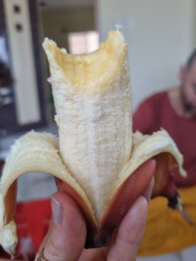 A partially peeled red banana is held in someone's hand. The banana peel is a reddish-brown color. The fruit inside is yellow and creamy. A person is partially visible in the background, wearing a maroon shirt. The background includes a room with a cabinet. The image appears to have been taken indoors. The location is unknown.