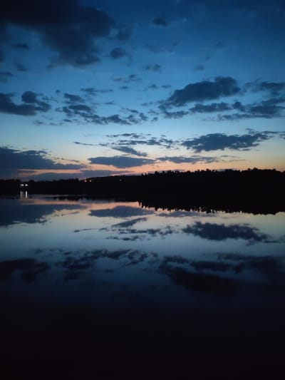 The image depicts a tranquil body of water, likely a lake or a wide river, reflecting a twilight sky. The sky is a mix of blues and grays, with scattered clouds. A dark, silhouetted treeline stretches along the horizon, possibly indicating a forest or dense vegetation. There are faint lights visible in the distance, possibly from buildings or street lamps, suggesting a town or settlement in the vicinity. The dark foreground suggests the vantage point is close to the water's edge. The image was probably taken at Ermolino, Russia, but there is no evidence of where in Ermolino it was taken.