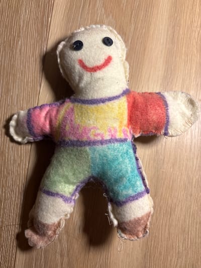 A homemade stuffed doll lies on a wooden floor. The doll is made of white fabric and has two dark blue buttons for eyes and a red stitched smile. Its torso is a patchwork of colorful felt, with purple stitching outlining the sections. The doll's arms and legs are also made of white fabric, with some sections of felt patchwork. The doll's feet are a brown and pink felt patch. The doll is positioned face up, with its arms and legs spread out. The wooden floor has a light brown grain. No specific location in Cesena, Italy is identifiable in the image.