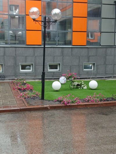 The image shows an outdoor decorative scene in front of a modern building with an orange and grey facade. A black lamppost with two spherical glass light fixtures stands in the center. The ground is covered with artificial green turf, bordered by red brick pavers and a planter with pink flowers. There are three large white spherical decorations placed on the turf, and a small decorative bicycle with a pot of flowers. The building has multiple windows, some of which reflect the surroundings. The pavement in the foreground is wet, indicating recent rain. This scene is located in the city of Kotino, Russia.