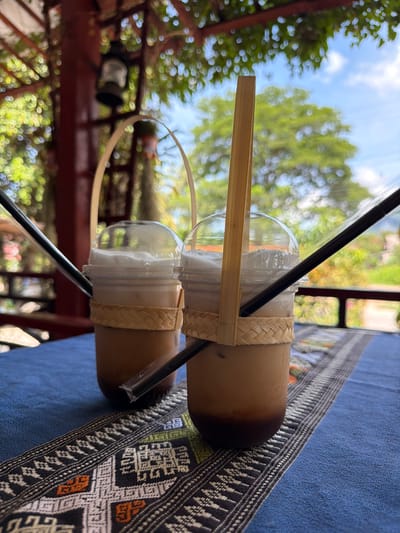 Two iced beverages are presented on a blue and patterned tablecloth in an outdoor setting, likely a cafe or restaurant in Vang Vieng, Laos. The drinks are in clear plastic cups topped with domes and contain a light brown liquid with a white foam layer. Each cup is adorned with a woven bamboo band and has a black straw inserted. A thin, flat wooden stick is also inserted into each cup, possibly serving as a handle or decorative element. The background is blurred, showing lush green foliage, a blue sky with white clouds, and hints of wooden structures, suggesting a tropical or semi-tropical environment. A vintage-style lantern is visible in the upper left corner, hanging from a wooden beam. The overall atmosphere is relaxed and informal, typical of a scenic tourist destination.