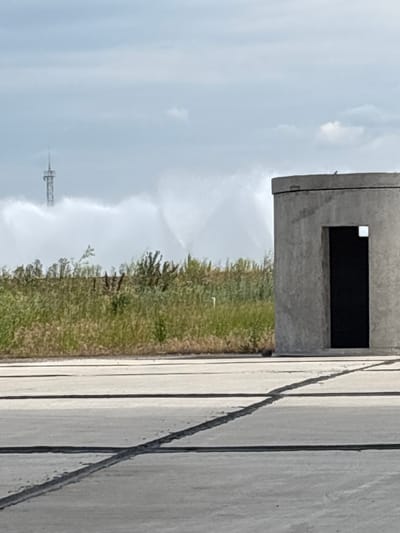 The image depicts an outdoor scene, likely an industrial or training area, given the presence of a concrete structure with a rectangular opening, suggestive of a firing range or observation post. This structure is situated next to a patch of overgrown grass and some greenery, possibly indicating a transition zone or undeveloped land. In the distance, a tall, slender structure resembling a communication tower or a part of a larger industrial complex is visible against a hazy sky. The ground in the foreground is paved with concrete slabs, marked by dark lines, suggesting a road or an airfield. The sky appears overcast with patches of light, hinting at a diffused daylight, possibly morning or late afternoon, with moderate weather. There is a fine mist or spray of water visible, arcing through the air from right to left, possibly from a sprinkler or a hose, suggesting some form of maintenance, training, or a cleaning activity. The overall mood is functional and somewhat stark, with no visible people or clear indicators of specific emotions. The image is oriented vertically, with the concrete structure and grassy area occupying the upper and middle sections, and the paved ground in the lower right. The location context is identified as Pivdennoukrainsk, Ukraine, which aligns with the utilitarian and somewhat industrial aesthetic. No text is visible in the image.