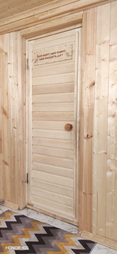 Here is a description of the image:

The image shows a wooden door inside what appears to be a sauna or bathhouse, given the text on the sign above the door. The door is made of horizontal wooden planks and has a round wooden doorknob. A sign at the top of the door has text in Russian: "Баня парит, баня правит, баня молодость даёт", which translates to "Bath steams, bath rules, bath gives youth". The surrounding walls are also made of wooden planks. The bottom of the image shows part of a rug with a chevron pattern in gray, black, and yellow. The location information indicates the photo was taken in Беркуты, Russia. There are no people visible in the scene.