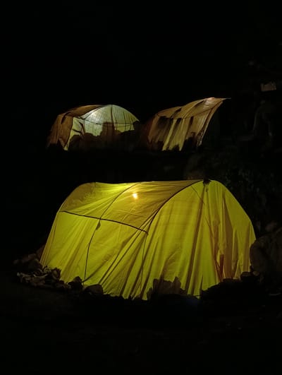 The image depicts two camping tents in Palchan, India, illuminated by artificial light sources within them at night. The foreground is dominated by a large, bright yellow tent, its fabric taut and curved, with a visible light bulb inside casting a warm glow. Rocks of varying sizes surround the base of this tent, suggesting a natural or somewhat rugged environment. In the background, higher and further back, a second tent, appearing lighter in color and more translucent, is also illuminated, revealing a subtle grid pattern within its fabric. The surrounding environment is dark, indicating nighttime, with the tents being the primary sources of light and visual interest. The scene appears to be set outdoors, possibly in a campsite or a natural area near Palchan.