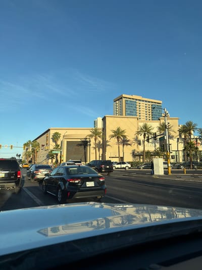 This media file captures a sunny, daytime street scene in Las Vegas, United States of America, viewed from inside a vehicle. The perspective is slightly rotated, showing an urban landscape filled with buildings, palm trees, and traffic.
**Subjects:** The central subjects are large, light-colored buildings, most notably the Sahara Las Vegas hotel, which features a distinctive façade and modern glass sections. Numerous palm trees line the street and stand in front of the buildings. The multi-lane road is packed with cars, including black sedans, SUVs, and vans, forming bumper-to-bumper traffic. Traffic lights and street signs are visible, guiding the flow of vehicles. The sky is a clear, bright blue with a faint crescent moon visible in the upper left. In the foreground, a highly reflective surface, likely part of the car's hood or dashboard, shows mirrored reflections of the buildings, palm trees, and sky.
**Activity or Event:** The primary activity is vehicular travel and queuing in traffic on a busy city street.
**Notable Details:** The time of day is clearly daytime, characterized by bright sunlight and a cloudless blue sky. The angle of the shadows suggests either morning or late afternoon. Location cues are abundant, with the prominent "SAHARA" sign confirming the Sahara Las Vegas hotel and resort. Nevada license plates are visible on several cars, further pinpointing the location. The general atmosphere is bustling and urban, typical of a major city like Las Vegas.
**Visible Text:** The most prominent text is "SAHARA" emblazoned on the hotel building, with "LAS VEGAS" beneath it. Other texts visible on the building include "MARCH 10 TRUES", "HOT SEAT DRAWINGS", and "WEDNESDAYS". A green street sign clearly reads "PARADISE RD", and another, partially obscured, appears to indicate "SAHARA AVE". A Nevada license plate "N 0 7221" is visible on one car. Reflections on the car's surface show reversed text, including the word "SAHARA".