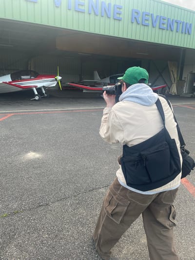An individual is depicted from behind, standing on a dark grey asphalt tarmac and photographing two light aircraft parked within a hangar. The location is identified by light blue raised letters spelling "VIENNE REVENTIN" across the light green corrugated metal facade of the hangar, indicating the Vienne Reventin Aerodrome near Chonas-l'Amballan, France.
The person wears a green baseball cap, a light grey hooded sweatshirt under a beige jacket, and brown cargo pants. A black shoulder bag with multiple zippered pockets hangs over their left shoulder. Both hands are holding a black digital camera, aimed forward at the aircraft.
Inside the open hangar bay, two single-engine propeller aircraft are visible. The aircraft on the left features a white fuselage with red stripes and a prominent yellow propeller. The aircraft on the right, partially obscured, also has a white fuselage with red stripes and displays the registration "F-GMXI" on its tail. The asphalt ground beneath the individual shows minor cracks and light-colored specks, with a red line marking visible on the right edge. The sky above is overcast.