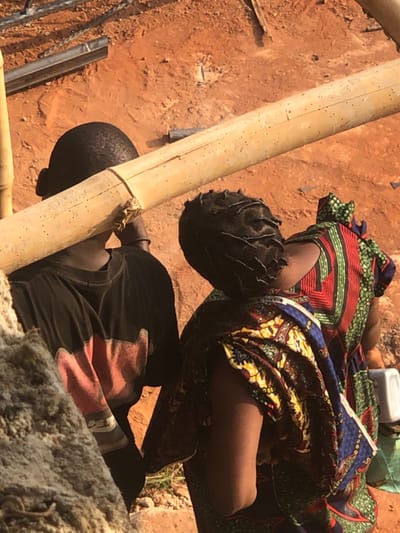 An aerial view captures two individuals on reddish-brown earth in Agukwu Nri, Nigeria. On the left, a person, likely male, with short dark hair, wears a dark t-shirt featuring a broad horizontal red band across the lower back. This individual carries a thick, light-colored bamboo pole horizontally across their right shoulder and upper back. A dark wristband is visible on their right arm. To the right, a second person, likely female, with dark hair styled in dreadlocks or braids, wears a vividly patterned dress or wrapper. The garment displays geometric designs in red, green, blue, yellow, and black. Her left arm is bent, revealing the inner elbow. In the upper left background, metallic bars or pipes rest on the ground. A blurry, light-colored object, possibly a rock, is in the lower left foreground. A small white container is partially visible on the far right. The scene depicts activity on unpaved terrain.