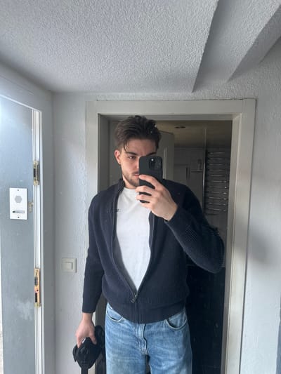 This image captures a young adult male taking a mirror selfie indoors in a residence located in Rafz, Switzerland. The male subject, standing roughly chest-up, has dark brown hair, styled with a part, and light facial stubble. He is wearing a white crew-neck t-shirt, an unzipped navy blue knitted cardigan with a front zipper, and light blue denim jeans. His right hand holds a dark-colored smartphone, positioned to capture his reflection. A black bag or pouch is partially visible in his left hand, held low near his left hip.

The immediate environment reflected in the mirror shows a white, textured ceiling overhead. To the left of the subject, a white door frame is visible, alongside a light-colored wall featuring a white rectangular intercom system with speaker perforations and two buttons. A gold-toned door strike plate is affixed to the door frame. To the right of the subject, beyond another white door frame, a reflection of an interior space, possibly a bathroom, reveals a multi-tiered metal towel rack or shelf unit. The lighting appears even, suggesting an indoor ambient light source.