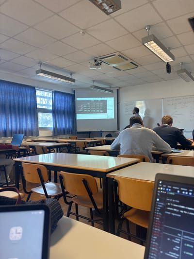 This image shows a classroom or lecture hall located in Porto Salvo, Portugal. The perspective is from a student's desk in the immediate foreground, where the top of a laptop screen displaying code or data and a tablet screen showing a clock application are visible.

In the mid-ground, several students are seated at individual desks with light-colored wooden tops and black metal frames, accompanied by matching chairs. Students are observed from behind; one wears a light grey long-sleeved shirt, and another wears a dark blue sweater. To the far left, a silver laptop is open on another desk.

At the front of the room, a male individual with dark hair and a beard stands in front of a white whiteboard, facing away from the camera, indicating a teaching or presentation role. The whiteboard contains handwritten mathematical equations and diagrams. Adjacent to the whiteboard, a large projection screen displays what appears to be a spreadsheet or a table of data.

The room is illuminated by natural light entering through two large windows on the left wall, covered by closed blue curtains, and by artificial light from several rectangular fluorescent fixtures mounted on the white suspended ceiling. The ceiling also features two recessed air conditioning vents and a single, circular smoke detector.
