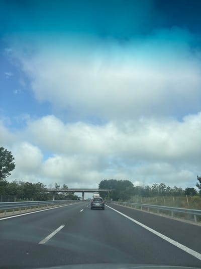 This media file captures a view from a moving vehicle on a multi-lane highway during the daytime, specifically in the Gorni Bogrov area of Bulgaria. The image is oriented vertically but depicts a horizontally stretching road.
The scene shows a modern asphalt highway with clear white lane markings. The vehicle from which the picture is taken appears to be in the rightmost lane. Further ahead, a dark-colored car, possibly a hatchback or small SUV, is visible in the same lane. The highway is bordered by a metal guardrail on the left, behind which are dense green trees and other foliage, suggesting a semi-rural or natural environment alongside the road. In the distance, an overhead structure, likely an overpass or bridge made of concrete or metal, spans across the highway.
The sky occupies the upper portion of the image. It is partly cloudy, with scattered white cumulus clouds against a varying blue background. There's a noticeable bright area in the upper left of the sky, indicating direct sunlight or a strong glare. The overall weather appears clear and bright.
No discernible people are visible, and no legible text can be identified on signs, vehicles, or other elements in the image. The activity taking place is typical highway travel.