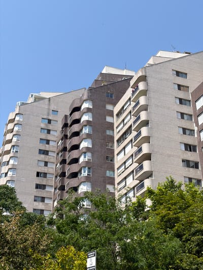 This media file features a low-angle shot of a Brutalist-style apartment building, likely in Courbevoie, France. The building's facade is characterized by its rough, textured concrete, geometric design, and numerous recessed balconies, creating a striking interplay of light and shadow. The architecture exhibits a repetitive pattern of windows, many of which are open or have blinds partially drawn, suggesting the building is inhabited. Lush green trees with dense foliage partially obscure the building on the right side of the frame, indicating a warm season. The sky is a clear, bright blue, implying it is daytime with good weather.

The image is vertically oriented, giving a sense of the building's immense height. The composition, with the building dominating the frame and reaching towards the sky, evokes a feeling of scale and urban density. There are no people visible, but the open windows and the presence of laundry hanging on a balcony hint at daily life within. A small, black and white sign with text is partially visible amongst the trees on the far right, though the text itself is illegible at this resolution. The overall impression is one of a modernist urban landscape bathed in natural light.