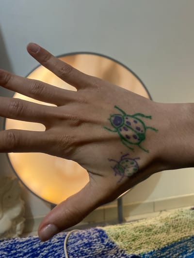 A hand with two drawn ladybugs is displayed prominently. The larger ladybug is drawn in green with dark purple spots and head, while the smaller one is drawn in purple with dark purple spots. The hand is fair-skinned with visible veins and wrinkles. The background features a light-colored surface on the left and a round, illuminated object behind the hand. To the right, a blue and green knitted item is partially visible. There is no information in the image to determine that the image was taken in Aubervilliers, France.