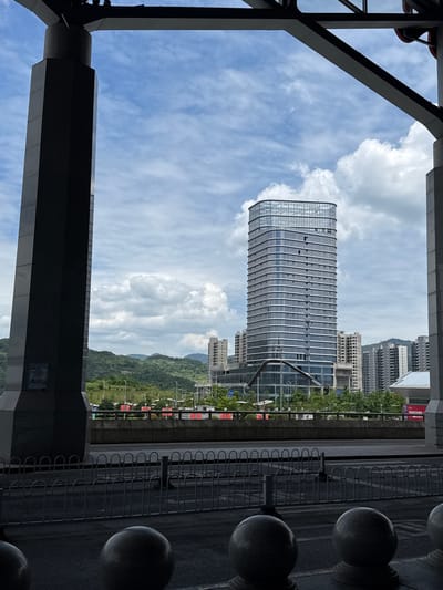 The image, taken in Yongding District, China, presents a view framed by a dark, structural steel or concrete framework, possibly a bridge or elevated walkway. The focus is a tall, rectangular glass-clad building, presumably an office or apartment tower, rising against a bright, partly cloudy sky. Behind the tower, a hillside covered in trees can be seen, with additional buildings in the distance. To the right, an ornamental iron railing runs alongside what appears to be a sidewalk or walkway, punctuated by spherical stone or concrete posts. No people are visible. The composition emphasizes the contrast between the modern architecture and the natural landscape, as well as the framing effect of the foreground structure.