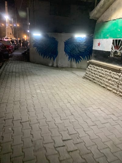 The image depicts a nighttime urban scene in Ma'raba, Syria. A paved walkway of interlocking grey concrete blocks occupies the foreground, extending into the middle distance. To the right, a building wall features a mural of large, blue and black stylized wings, illuminated by two rectangular LED lights mounted directly above. Adjacent to this, the wall transitions to another section painted with the Syrian opposition flag: horizontal stripes of green, white (with a red star), and black. Below the flag, the wall is constructed from light-colored, rough-hewn stone blocks, and a small, barred window with radial spokes is visible. To the left of the paved walkway, several parked cars, including dark and red sedans, line a narrow street. Beyond the vehicles, a group of indistinct individuals is visible, congregating near a tall, light-colored monument or obelisk illuminated by streetlights in the background. The overall environment is dimly lit by artificial light sources.
