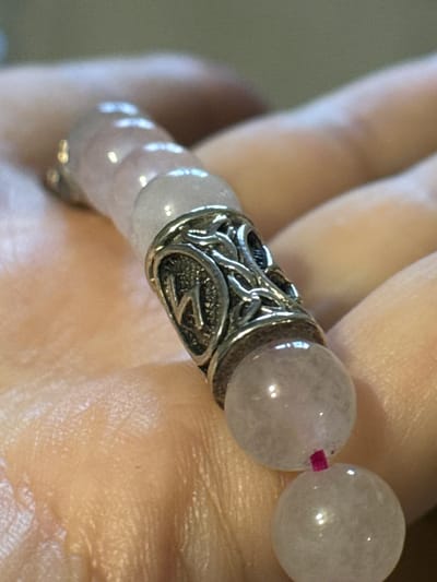 A close-up image shows a pink beaded bracelet with a silver ornate cylindrical spacer held in a human hand. The beads appear to be rose quartz. A pink cord threads between two beads. The silver spacer has a textured surface with a letter 'Z' design on one side. The skin of the hand is visible with pores and wrinkles, suggesting an older person. No details indicate the location of Novo-Nikol'skoe, Russia.
