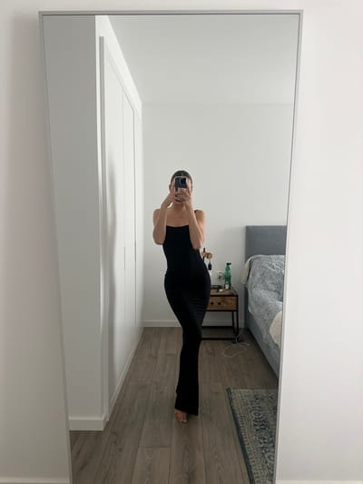 A full-length mirror selfie captures an individual standing barefoot in a room in Ericeira, Portugal. The person, appearing female, wears a black, form-fitting, spaghetti-strap maxi dress. They are actively holding a dark-colored smartphone to photograph their reflection, obscuring their face.
The room features white walls and light-colored wooden floorboards. On the left side of the reflection, white built-in wardrobes or closet doors are visible. On the right, a bed with a grey fabric headboard and a patterned grey and white duvet is present, alongside a small, dark-patterned rug. A minimalist nightstand, constructed from dark metal and wood, stands between the individual and the bed. Items on the nightstand include a clear glass bottle, a green plastic bottle, a black electronic device, and various associated cables.