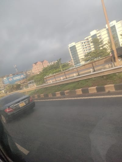 The image depicts an urban street scene under an overcast sky, located in Theta ward, Kenya. In the foreground, the lower left quadrant shows a partial view of a vehicle's dark interior frame. The mid-ground features a dark asphalt road with a partial white lane marking on the left. A black Mercedes-Benz C180 sedan, bearing Kenyan license plate KUB 876G, is visible on the road ahead, angled slightly to the right. To the right of the road is a raised concrete median, painted with alternating black and light brown blocks, topped by a strip of grass and a metallic guardrail.

In the background, beyond the median and guardrail, an elevated road or overpass is visible with a vehicle partially seen on its right lane. Lush green trees and vegetation line the area behind the elevated structure. Several buildings are present, most notably a tall, multi-story building with light-colored facades and blue-tinted windows, featuring the "NISS" logo at its summit. Adjacent to it, another similar building displays "KISS" signage. Further to the left and rear, multiple smaller buildings with reddish-brown tiled roofs and lighter facades are visible. A large blue billboard featuring the Pepsi logo and the text "THIRSTY PREMIUM" is prominent on the left, partially obscured by a tall communications tower. No individuals are distinctly visible.