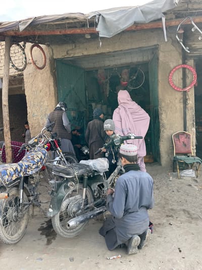 This image depicts an outdoor scene in Nakhonay, Afghanistan, in front of a rudimentary building functioning as a motorcycle repair shop. The structure features rough, unpainted mud or concrete walls and a corrugated metal green door, which is open. A makeshift roof, constructed from tarpaulin and various materials, covers the entrance.

Multiple individuals are present. In the foreground, a young male, wearing a gray tunic, black neck scarf, and a white crocheted skullcap, kneels on the dusty ground, facing away, engaged in repairing a dark-colored motorcycle. Adjacent to him, a child in a gray beanie with "Selfie" text and a dark jacket stands near the handlebars of the same motorcycle. To the right, a person in a long, light-pink garment stands with their back to the viewer, facing into the shop entrance. Further left, another person in a dark vest over a gray shirt and a patterned headscarf stands near the shop, while a person in dark brown attire and a beanie is partially visible inside the shop's entry. Another child is faintly visible peering from an opening to the far left.

Two motorcycles are prominent in the mid-ground. The one on the left has a red frame and a blue camouflage-patterned seat. The motorcycle being worked on is dark green or black, partially disassembled, with its seat covered in clear plastic. The ground is dirt, with some debris including a visible discarded syringe near the kneeling person. A broken, red-upholstered chair with a metal frame leans against the wall on the right. Various circular objects, appearing to be parts or tires, hang from the building's exterior, including a red-wrapped ring on the right and two white-wrapped rings on the left. The sky is overcast, providing diffuse natural lighting.