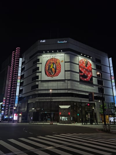 At night, a view of a building in Shinjuku, Japan, featuring prominent advertising for Louis Vuitton. The building is multi-tiered and well-lit, adorned with illuminated Louis Vuitton advertisements. The street is quiet, with a crosswalk marked by broad white stripes intersecting the foreground. Street lights and signage are visible along the road, contributing to the urban atmosphere. A building with pink neon lighting is near the bottom of the image. The name of the building, "Frente," is displayed prominently.