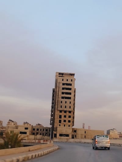 The scene, located in Al-Qadam Municipality, Syria, depicts an urban environment under a light-colored sky with a subtle blue-to-peach gradient, indicative of dawn or dusk. Dominating the central-right midground is a tall, multi-story concrete building, appearing either unfinished or extensively damaged, featuring numerous empty rectangular apertures and visible rebar. To its left and receding into the background, several additional concrete structures, predominantly lower-rise, also display signs of incompleteness or damage. A two-lane asphalt road, bordered by concrete barriers, curves from the left foreground towards the right midground. A silver passenger van is observed driving away from the viewer on this road. Blurred green foliage is present in the far-left foreground. No human figures are visible.