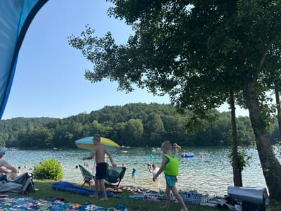 The image depicts a scene at a lake, likely a recreational area in Blankenheim, Germany, on a sunny day. In the foreground, a blue tent is partially visible on the left, framing the view. A person, likely a child, wearing a green life vest and striped swim trunks, is walking towards the water from the right. Another child, bare-chested with dark swim trunks, is standing near a beach chair. A colorful umbrella provides shade. The lake is populated with numerous people swimming, floating on inflatables, and enjoying water activities. In the background, a densely wooded hillside overlooks the lake, with lush green trees covering the slopes under a clear blue sky. Picnic blankets and bags are spread out on the grassy shore in the foreground, suggesting a day trip or picnic.