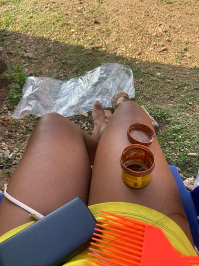 The image shows a close-up view of a person's legs, likely outdoors in a natural setting. The ground appears to be a mix of dirt and sparse green vegetation. To the left, a crumpled piece of clear plastic sheeting is visible.

On the person's thigh, there is an open yellow tin, possibly containing some kind of product. The lid of the tin is placed next to it. Next to the tin is an orange plastic comb with thick tines, which appears to have some hair tangled in it. Also on the thigh, resting against the person's leg, is a dark blue rectangular object that resembles a power bank, with a white cable peeking out from underneath. The person is wearing bright yellow clothing.

In the background, there is a hint of a blue surface, possibly a chair or seating. The lighting suggests it is daytime, with shadows indicating sunlight. There is no explicit indication of the weather, but the presence of greenery suggests it is not a dry season. The overall impression is a casual, outdoor setting, potentially a place where someone might be tending to their hair. There is no visible text that can be clearly read.