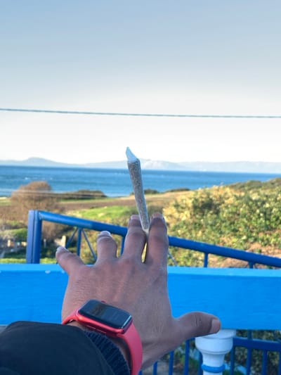 A hand holds a lit cigarette or joint, positioned horizontally. The hand is adorned with a red smartwatch. Background details include a blue structure, possibly a railing or fence, a body of water extending to a distant landmass or coastline. The sky above is overcast. The overall scene suggests an outdoor setting, perhaps a coastal area.