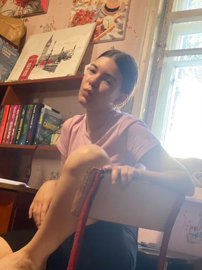 A young woman with dark hair pulled back sits in a chair, looking at the camera with an unexpressive gaze. She is wearing a pink t-shirt and black shorts. Her right arm rests on the back of the chair, while her left arm is bent and her hand is resting on her knee. Behind her, a bookshelf is filled with books. On the wall above the bookshelf are several framed pictures, including one of Big Ben and a red telephone booth. A window is visible to the right of the woman, letting in natural light. The room appears to be a personal space, possibly a bedroom or living area, with a casual and somewhat cluttered atmosphere.