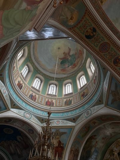 The image shows the interior of a domed building, likely a church or cathedral, with elaborate frescoes and architectural details. The viewpoint is looking upwards towards the dome.
**Dome and Ceiling:** The central dome features a painting of a religious figure descending from a bright light, surrounded by clouds. The dome itself is painted in shades of blue and light green, with some areas appearing to represent the sky. Surrounding the base of the dome is a band of windows, arch-shaped and divided into panes by white mullions. Below the windows, a circular frieze displays a series of painted religious figures in square or rectangular panels, separated by decorative motifs. Faint inscriptions in Cyrillic script are visible on a turquoise band encircling the dome. The ceiling also incorporates ornate painted borders and decorative patterns.
**Architecture and Decoration:** Arches and vaulted ceilings are visible in the periphery, also adorned with frescoes. These paintings depict various religious scenes and figures, including winged beings and robed individuals. The color palette is rich, with greens, blues, reds, and gold tones used throughout the artwork.
**Objects:** A large, ornate brass chandelier with numerous candle holders hangs from the center of the dome, suspended by a thin cable.
**Environment:** The lighting suggests an indoor environment, with light entering through the windows and contributing to the illuminated effect on the ceiling. The overall impression is one of grandeur and spiritual significance, characteristic of religious architecture.
**Location:** This interior is identified as being located in the city of Nogornyi, Russia.