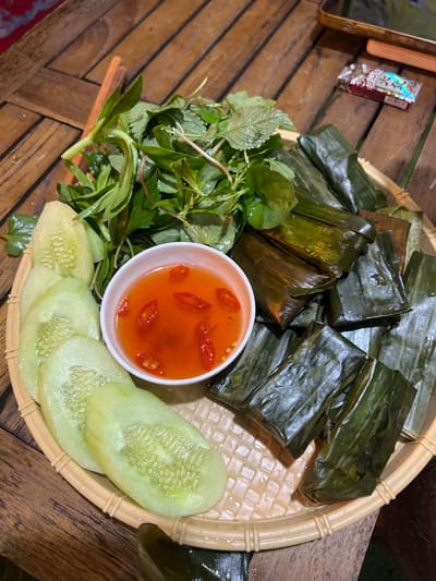 In Bắc Hà, Vietnam, a woven bamboo tray rests on a wooden table. The tray holds a collection of food items: a pile of fresh green herbs, several slices of cucumber, a small white bowl filled with a reddish dipping sauce containing slices of red chili, and a stack of rectangular food parcels wrapped in dark green leaves. The overall impression is of a rustic, outdoor dining setting.