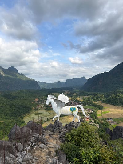 A white statue of Pegasus, the winged horse from Greek mythology, stands prominently on a rocky outcrop overlooking a vast valley in Naka, Laos. The statue is positioned on the right side of the frame, facing left, with its wings spread. A stylized blue and yellow emblem is visible on its hindquarters. The foreground is dominated by rugged grey rocks and some sparse vegetation, with a rusted wire fence running across the left side.

The middle ground reveals a sprawling rural landscape. Green rice paddies, some flooded and reflecting the sky, are interspersed with patches of dry, harvested fields. A small village with houses and a road winding through it is nestled within the valley. Beyond the village, lush green forests cover the lower slopes of dramatic, mist-shrouded mountains that form the backdrop of the scene. The sky above is a dramatic mix of blue patches and heavy, grey clouds, suggesting dynamic weather conditions. The overall impression is one of majestic natural beauty enhanced by the striking mythological statue.