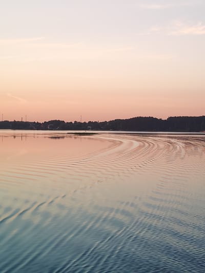 The image shows a tranquil body of water reflecting the soft colors of a sunset. The sky is a gradient of pale pink and light blue, with a few wispy clouds. On the horizon, a dark line of trees and some distant buildings with tall, slender structures that resemble communication towers are visible. The water's surface is disturbed by gentle ripples, creating a pattern of light and shadow that extends from the right side of the frame. The overall impression is one of peacefulness and natural beauty. The location is identified as Terekhovo, Russia.