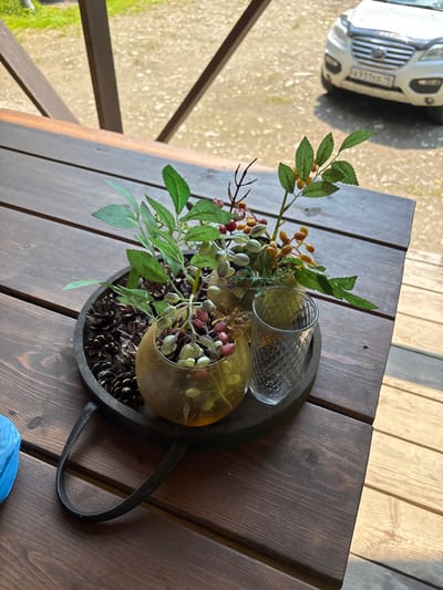 An arrangement of artificial foliage and berries sits in a shallow, round, dark-colored tray filled with pinecones on a dark wooden table. The arrangement includes green leaves, small red and yellow berries, and light green buds. A clear, patterned glass is placed to the right of the arrangement. In the background, a white vehicle with a license plate is visible. The scene appears to be outdoors, possibly on a porch or patio. The location is identified as Bol'shaya Rechka, Russia.