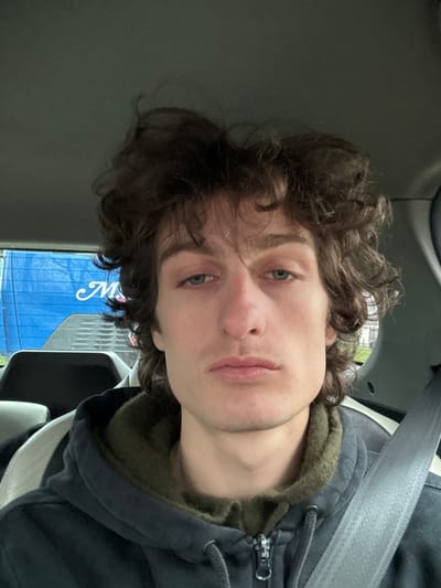 A young adult male is seated inside a vehicle, depicted in a head-and-shoulders portrait. He has light skin, blue eyes, a prominent nose, and dark brown, wavy hair. He wears a dark, possibly navy or black, zippered hoodie over an olive-green hooded garment. A grey seatbelt is visible across his chest, extending from his right shoulder diagonally downwards. The vehicle's interior is dark-toned, with a grey and white patterned headrest visible behind him to his left. Through the rear window of the vehicle, a large blue surface with partial white lettering, appearing to be an 'M' and other indistinct characters, is visible, suggesting another vehicle or a building with signage. Faint outlines of trees or foliage and a light-colored structure are also discernible in the background. The scene is located in Alfortville, France.