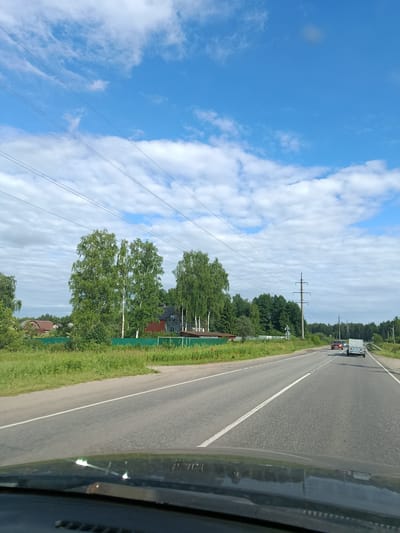 The image depicts a highway scene in Тополиный, Russia, on a sunny day with scattered clouds. The perspective is from inside a vehicle, looking forward along the asphalt road. The road has white dashed lines dividing the lanes and a solid white line on the right edge.
On the left side of the road, there is a grassy verge with a green fence. Behind the fence, several houses with brown roofs are visible, interspersed with lush green trees. Prominent in this area are tall birch trees with distinctive white bark and dense green foliage. Power lines stretch across the sky, supported by tall utility poles.
On the right side of the road, the landscape is more wooded, with a dense line of trees. Several vehicles are traveling on the highway. Ahead, a red car is visible, followed by a white trailer being towed by another vehicle. A power line pole is also visible on the right.
The sky is a vibrant blue with fluffy white clouds, suggesting fair weather. The time of day appears to be daytime, likely late morning or early afternoon, given the brightness of the sun and the shadows. The scene conveys a sense of travel and a rural or semi-rural setting. No visible text is present in the image.