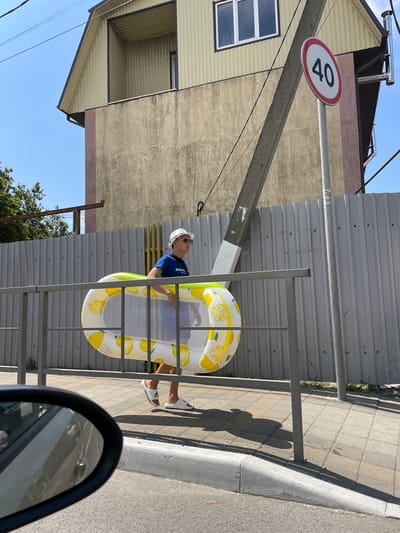 A young person wearing a white bucket hat, sunglasses, a blue t-shirt, and white slide sandals walks along a sidewalk carrying a yellow and white lemon-patterned inflatable pool float. The person is walking past a grey corrugated metal fence and a metal railing. In the background, a building with a beige facade and a yellow-paneled upper section is visible, along with a speed limit sign indicating 40. The scene appears to be in Dzhubga, Russia, given the context. The sun is shining, casting shadows on the ground, and the sky is clear and blue. A car's side mirror is visible in the lower left corner of the frame.
