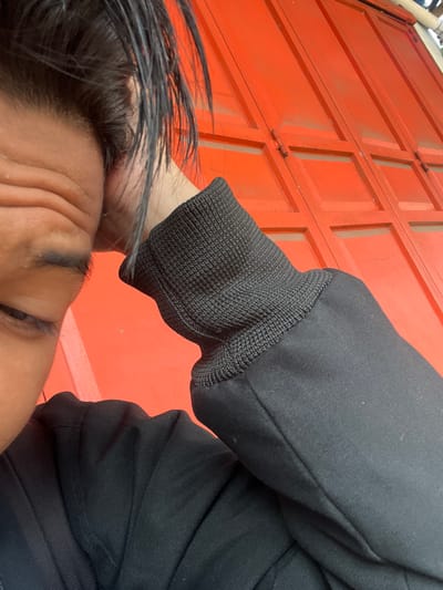 A close-up, low-angle shot shows a person's face and arm in the foreground, with a bright orange, paneled background. The person's dark hair is wet and styled messily, framing the left side of their face. Their eyes are partially closed, and their forehead is furrowed, suggesting a relaxed or perhaps tired expression. The person is wearing a black jacket with a ribbed cuff on their right arm. The background consists of large, interlocking orange panels that resemble garage doors or shutters, with visible hinges and seams. The bright orange color and industrial appearance of the background suggest an outdoor or utilitarian setting. There are no visible signs of time of day or weather conditions. The image does not contain any discernible text. The overall impression is a casual, candid moment captured from a unique perspective.