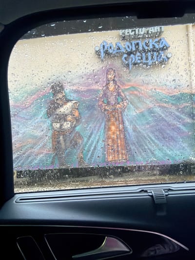 The image shows a view through a car window, likely during a car wash. The window is covered in water droplets, obscuring the view. On the other side of the window, there is a building with a mural. The mural appears to depict two figures, possibly people, in a stylized or artistic fashion. The colors in the mural are a mix of blues, purples, and browns. There is also a sign that is mostly obscured, but the word "Restaurant" is visible and the place is called "Podkova St.", likely referencing the restaurant name and address within the city of Davidkovo, Bulgaria. The interior of the car is visible, including a car door panel.