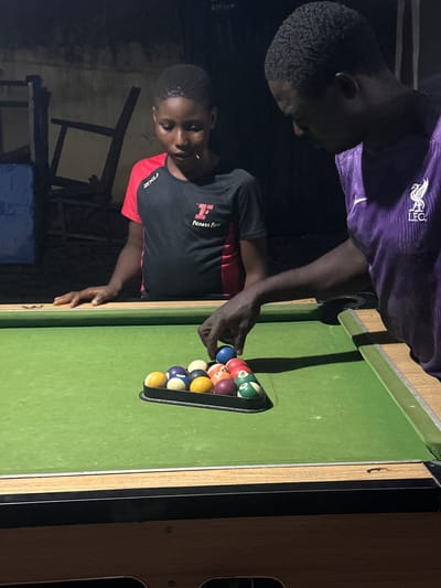 In an outdoor setting, two African men are positioned at a pool table in Ogwashi-Uku, Nigeria. One man, wearing a purple shirt with "LFC" visible, leans over the table. The other man, dressed in a black and red athletic shirt, stands beside the table, arranging the pool balls in a triangular rack. The pool table is green, with a set of multi-colored pool balls in the rack. The setting appears to be at night. A wooden chair and a blue container are visible in the background.