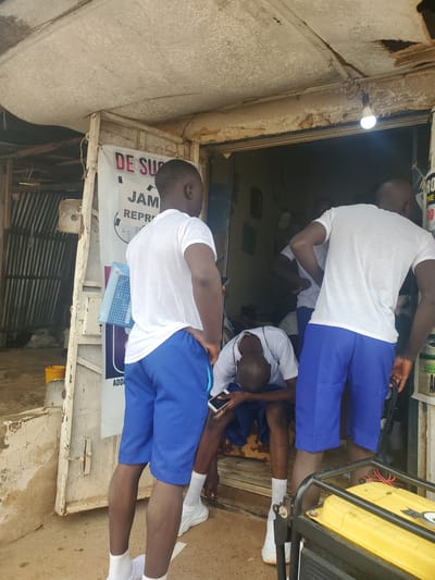 A group of young Black men in matching white t-shirts and blue shorts stand outside a storefront. One man in the foreground is facing away from the camera, his left hand on his hip. Another man is seated with his head down, looking at his phone. The storefront is weathered and appears to be made of corrugated metal and wood. A sign with African script hangs on the left side of the store. A yellow generator is partially visible in the lower right corner of the image. The scene is dimly lit, with a single light bulb hanging from the ceiling of the storefront. The ground is dirt and littered with debris.